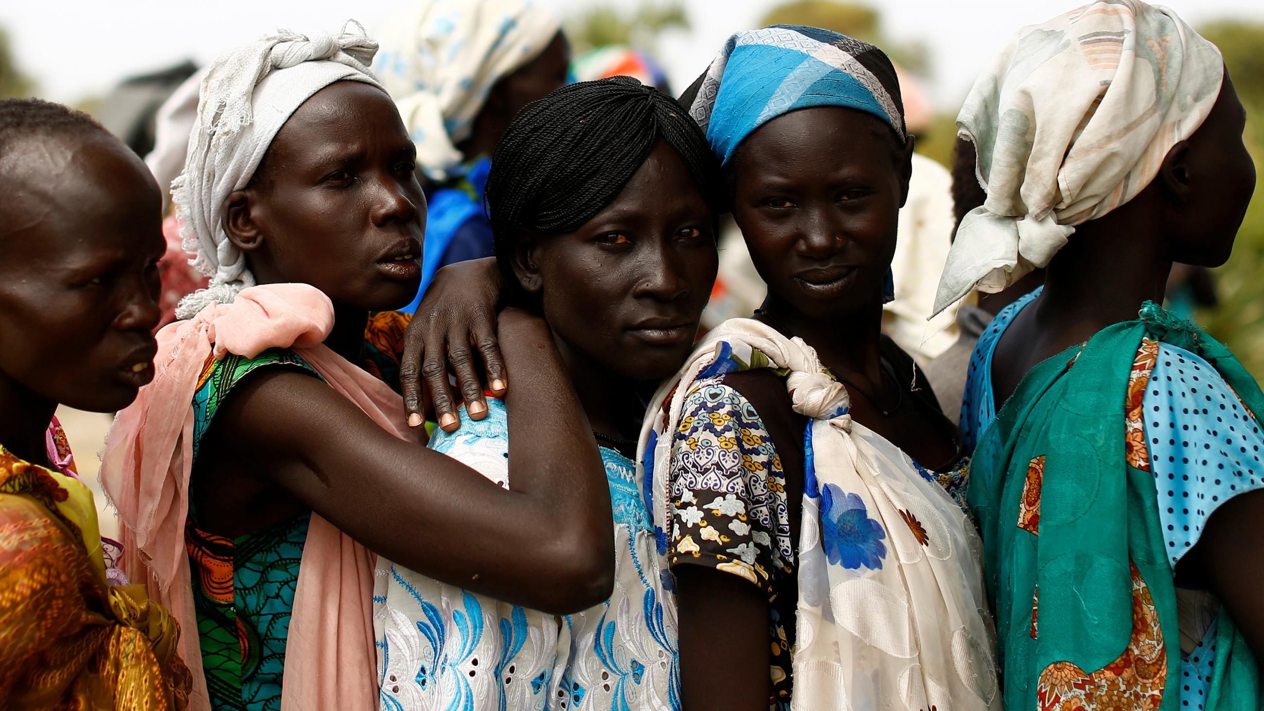 Picture shows five women in a line with more lined up on the other side of them. The three women in the middle are standing very close, and the third has her arms around the shoulders of the second, suggesting they all know each other. This photo is moving in its simple, straightforward humanity.