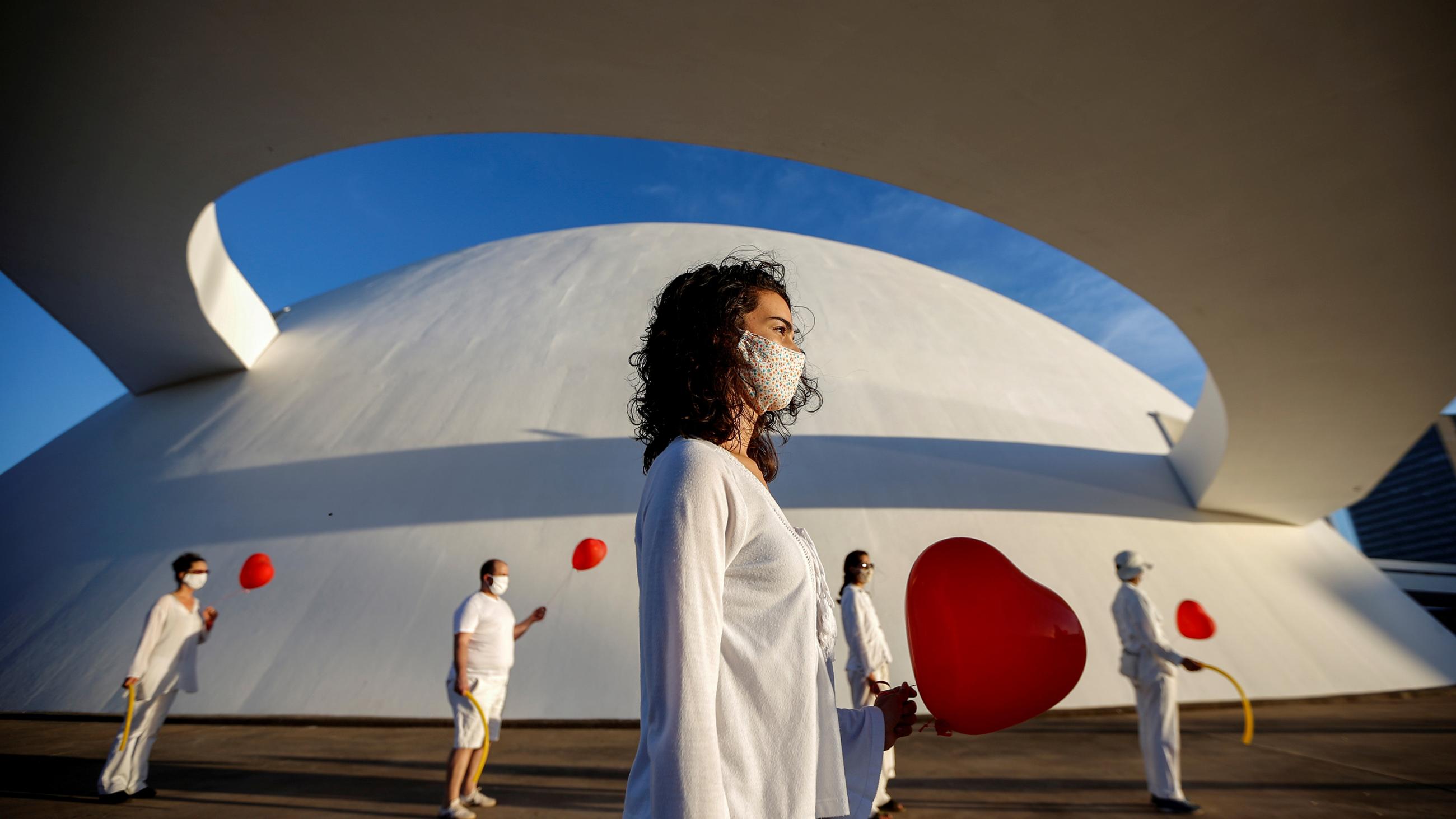 The photo shows a performance art piece with a woman wearing white in the foreground and four people wearing white and holding red balloons in the background.